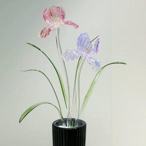 Op de afbeelding: Een hoge, zwarte glazen vaas met verticale ribbels die twee glazen bloemen bevat. De ene bloem is roze met een doorschijnende textuur en de andere is lichtpaars. Beide bloemen hebben lange, heldere glazen stelen en dunne, groene bladeren.
