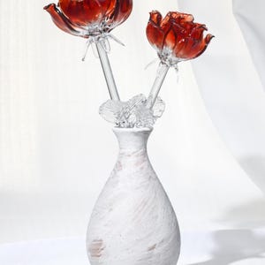 May include: Two glass roses with deep red petals and clear stems are arranged in a white ceramic vase. The vase has a textured, brush-stroke finish with subtle pinkish undertones. Clear glass leaves are attached to the stems near the vase opening.