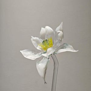 May include: A handcrafted glass flower sculpture featuring translucent white petals with delicate, wavy edges. The center of the flower displays bright yellow stamens and a green pistil. The entire piece is supported by a clear glass stem.