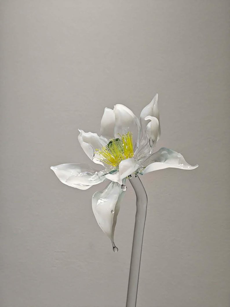 May include: A handcrafted glass flower sculpture featuring translucent white petals with delicate, wavy edges. The center of the flower displays bright yellow stamens and a green pistil. The entire piece is supported by a clear glass stem.