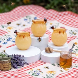 May include: Three small, yellow glass honey pots with dripped honey rims and bee accents sit on white pedestals. A small jar of honey, a honey dipper, and a tin of raw clover honey are also present. The scene is set on a red and white checkered picnic blanket.
