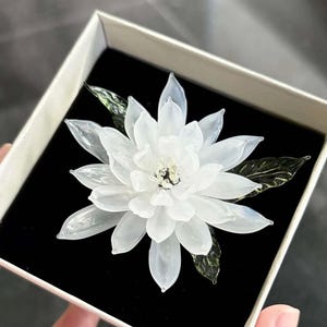 May include: A white glass flower brooch with translucent petals and dark green glass leaves. The flower features a central cluster of small, clear glass beads. It is presented inside a square box with a black velvet interior lining.