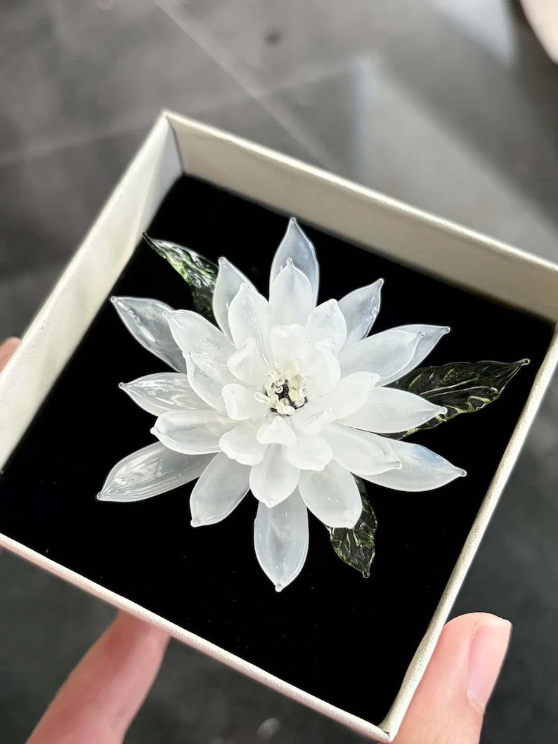 May include: A white glass flower brooch with translucent petals and dark green glass leaves. The flower features a central cluster of small, clear glass beads. It is presented inside a square box with a black velvet interior lining.