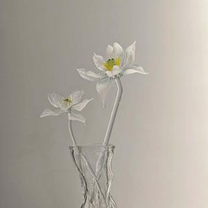 May include: Two white glass flowers with yellow centers and translucent petals are arranged in a clear, textured glass vase. The flowers have long, slender stems and are positioned upright, showcasing delicate craftsmanship and a smooth, glossy finish.