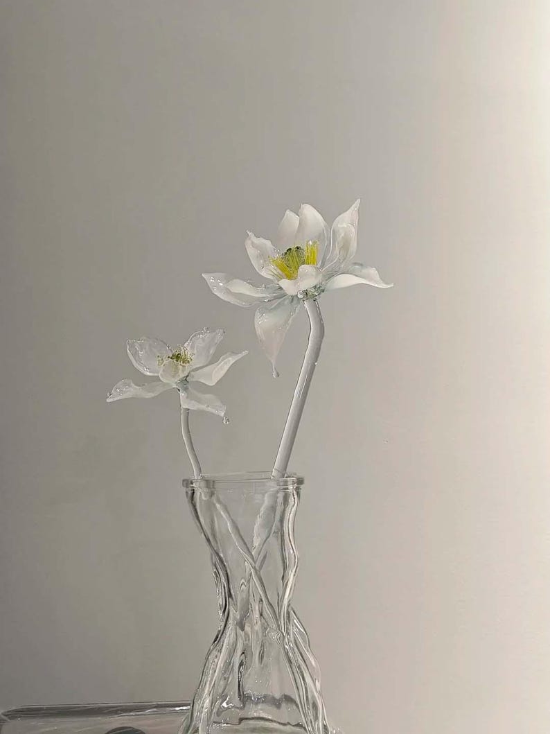 May include: Two white glass flowers with yellow centers and translucent petals are arranged in a clear, textured glass vase. The flowers have long, slender stems and are positioned upright, showcasing delicate craftsmanship and a smooth, glossy finish.