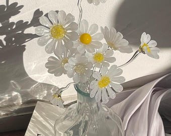 Könnte beinhalten: Eine klare Glasvase mit einem Strauß aus handgefertigten Glasgänseblümchen. Jede Blume hat zarte weiße Blütenblätter und eine leuchtend gelbe Mitte. Die Stiele sind dünn und transparent und in dem gerippten Glasgefäß angeordnet.