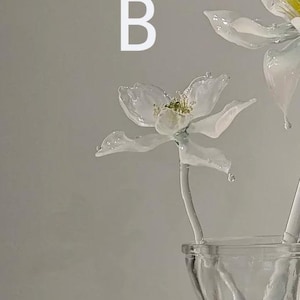 May include: A glass flower sculpture with white petals and a yellow center, featuring the letter B in white text above it. The flower is attached to a white stem and is positioned inside a clear glass vase with a textured, twisted design.