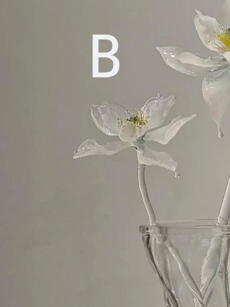 May include: A glass flower sculpture with white petals and a yellow center, featuring the letter B in white text above it. The flower is attached to a white stem and is positioned inside a clear glass vase with a textured, twisted design.