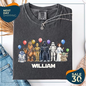 a t-shirt with a graphic design of various Star Wars characters, including Chewbacca, Darth Vader, and the Yoda from the Star Wars franchise, along with colorful balloons. The t-shirt is displayed on a white background, and there are jeans visible in the background.