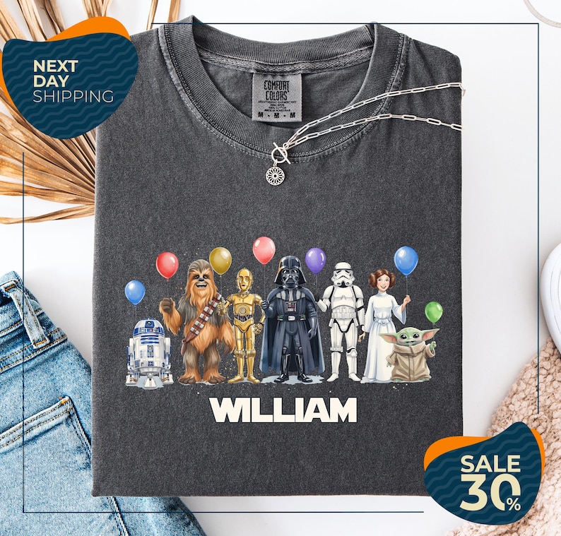 a t-shirt with a graphic design of various Star Wars characters, including Chewbacca, Darth Vader, and the Yoda from the Star Wars franchise, along with colorful balloons. The t-shirt is displayed on a white background, and there are jeans visible in the background.