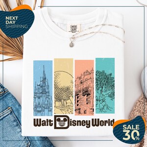a white t-shirt with a graphic design of Disney World landmarks, including a castle, an Eiffel Tower, and a roller coaster. The t-shirt is displayed on a white background, and there are jeans and a sweater visible in the background. The image also includes text that reads "Next Day Shipping" and "SALE 30%".