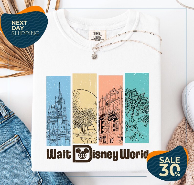 a white t-shirt with a graphic design of Disney World landmarks, including a castle, an Eiffel Tower, and a roller coaster. The t-shirt is displayed on a white background, and there are jeans and a sweater visible in the background. The image also includes text that reads "Next Day Shipping" and "SALE 30%".