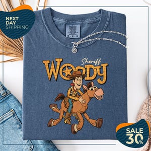 a blue t-shirt with a cartoon image of Woody, a character from the Toy Story movie, riding a brown horse. The t-shirt is displayed on a white background, and there are jeans and a sweater visible in the background.
