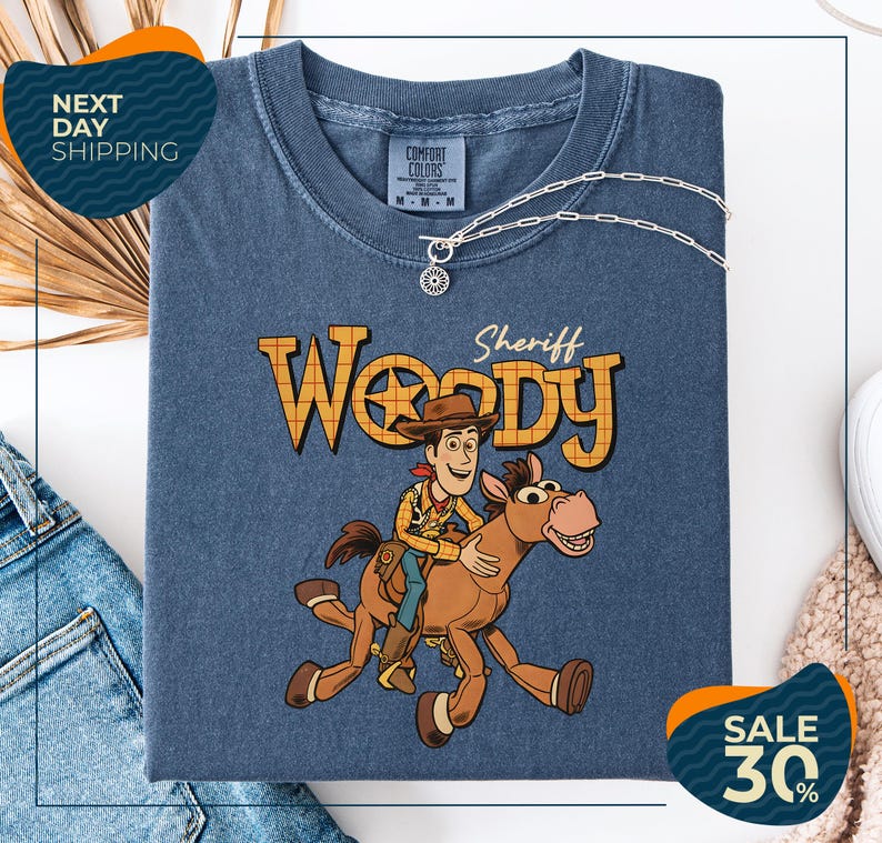 a blue t-shirt with a cartoon image of Woody, a character from the Toy Story movie, riding a brown horse. The t-shirt is displayed on a white background, and there are jeans and a sweater visible in the background.