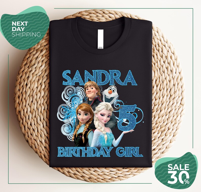 A black t-shirt with a design featuring characters from the Disney movie Frozen, specifically Elsa, Olaf, and Anna. The text on the t-shirt reads "SANDRA BIRTHDAY GIRL".