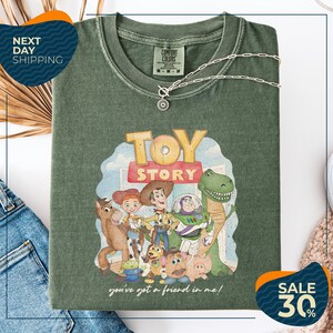 a green t-shirt with a cartoon character design, including a dinosaur, a cowboy, and other characters from the Toy Story franchise. The t-shirt is displayed on a white background, and there are jeans and a necklace visible in the foreground.