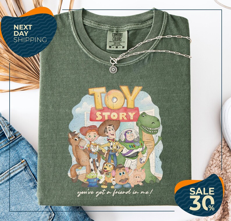 a green t-shirt with a cartoon character design, including a dinosaur, a cowboy, and other characters from the Toy Story franchise. The t-shirt is displayed on a white background, and there are jeans and a necklace visible in the foreground.