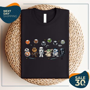 a black t-shirt with a graphic design of various Star Wars characters, including Star Wars, Darth Vader, and Baby Yoda, arranged in a row.