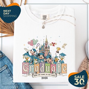 a white t-shirt with a colorful graphic design depicting Disney characters in graduation attire, along with a castle and other Disney-themed elements. The text on the t-shirt reads "Graduate from the Tasse to the Castle" and "2026". The t-shirt is displayed on a white background, with a pair of jeans and a sweater visible in the background. There are also two sale signs in the image, one indicating a 30% discount and the other promoting next day shipping.