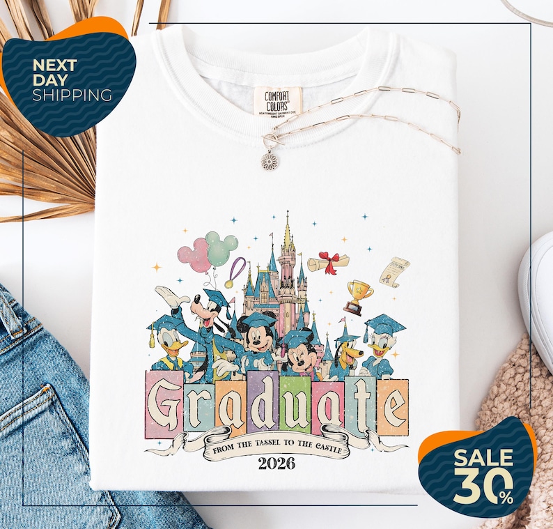 a white t-shirt with a colorful graphic design depicting Disney characters in graduation attire, along with a castle and other Disney-themed elements. The text on the t-shirt reads "Graduate from the Tasse to the Castle" and "2026". The t-shirt is displayed on a white background, with a pair of jeans and a sweater visible in the background. There are also two sale signs in the image, one indicating a 30% discount and the other promoting next day shipping.