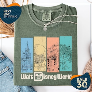 a green t-shirt with a graphic design of Disney World landmarks, including a castle, roller coaster, and other iconic structures. The t-shirt is displayed on a white background, and there are two sale tags visible, one for the t-shirt and one for a pair of jeans.