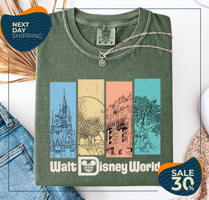 a green t-shirt with a graphic design of Disney World landmarks, including a castle, roller coaster, and other iconic structures. The t-shirt is displayed on a white background, and there are two sale tags visible, one for the t-shirt and one for a pair of jeans.