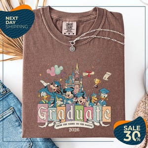 a brown t-shirt with a colorful graphic design depicting Disney characters in a graduation gown, surrounded by a castle and other decorative elements. The text on the t-shirt reads "Graduate from the Tassle to the Castle" and "2026". The t-shirt is displayed on a white background, with a pair of jeans and a sweater visible in the background.