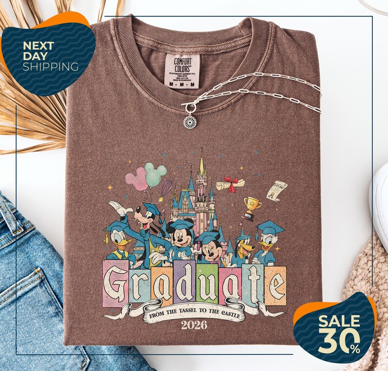 a brown t-shirt with a colorful graphic design depicting Disney characters in a graduation gown, surrounded by a castle and other decorative elements. The text on the t-shirt reads "Graduate from the Tassle to the Castle" and "2026". The t-shirt is displayed on a white background, with a pair of jeans and a sweater visible in the background.