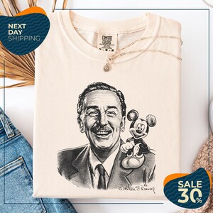 a t-shirt with a black and white illustration of Walt Disney, along with a necklace and a Mickey Mouse plush toy. The t-shirt is displayed on a white background, and there are some jeans and a sweater visible in the background.