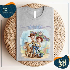 a gray t-shirt with a cartoon illustration of characters from the Toy Story franchise, including Woody, Buzz Lightyear, and an alien character. The t-shirt is displayed on a woven placemat, and there are two sale signs indicating a 30% discount on the item.