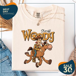 a white t-shirt with a cartoon image of Woody, a character from the Toy Story movie, riding a horse. The t-shirt is displayed on a white background, and there are some jeans and a sweater visible in the background.