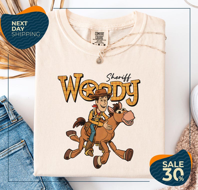 a white t-shirt with a cartoon image of Woody, a character from the Toy Story movie, riding a horse. The t-shirt is displayed on a white background, and there are some jeans and a sweater visible in the background.
