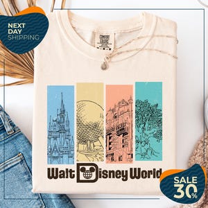 A white t-shirt with a graphic design featuring illustrations of various Disney landmarks, including the Eiffel Tower, the castle, and the roller coaster, along with the text "Walt Disney World". The t-shirt is displayed on a white background, and there are jeans and a sweater visible in the background.