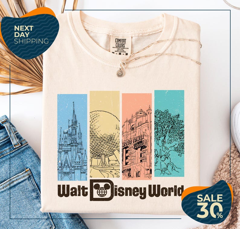A white t-shirt with a graphic design featuring illustrations of various Disney landmarks, including the Eiffel Tower, the castle, and the roller coaster, along with the text "Walt Disney World". The t-shirt is displayed on a white background, and there are jeans and a sweater visible in the background.