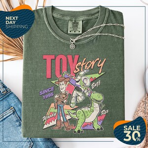 a green t-shirt with a colorful graphic of Woody and Buzz from the Toy Story movie, along with the text "Toy Story" and "Since 1995". The t-shirt is displayed on a white background, and there are jeans and a necklace visible in the foreground.