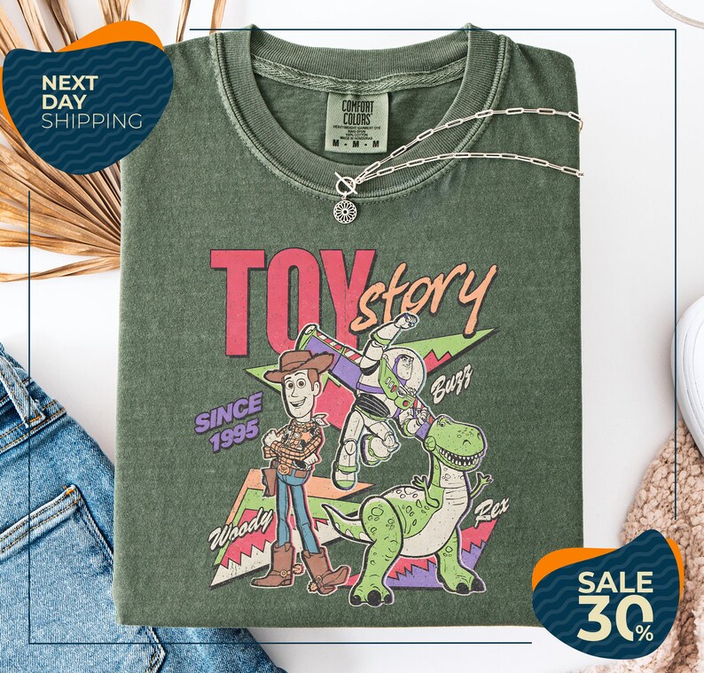 a green t-shirt with a colorful graphic of Woody and Buzz from the Toy Story movie, along with the text "Toy Story" and "Since 1995". The t-shirt is displayed on a white background, and there are jeans and a necklace visible in the foreground.