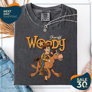 a gray t-shirt with a cartoon image of Woody, a character from the Toy Story movie, riding a brown horse. The t-shirt is displayed on a white background, and there are jeans and a necklace visible in the foreground.