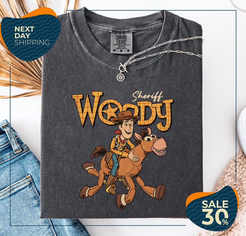 a gray t-shirt with a cartoon image of Woody, a character from the Toy Story movie, riding a brown horse. The t-shirt is displayed on a white background, and there are jeans and a necklace visible in the foreground.