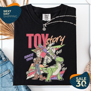 a black t-shirt with a colorful graphic design depicting characters from the Toy Story movie, including Woody and Buzz. The t-shirt is displayed on a white background, and there are jeans and a necklace visible in the foreground.
