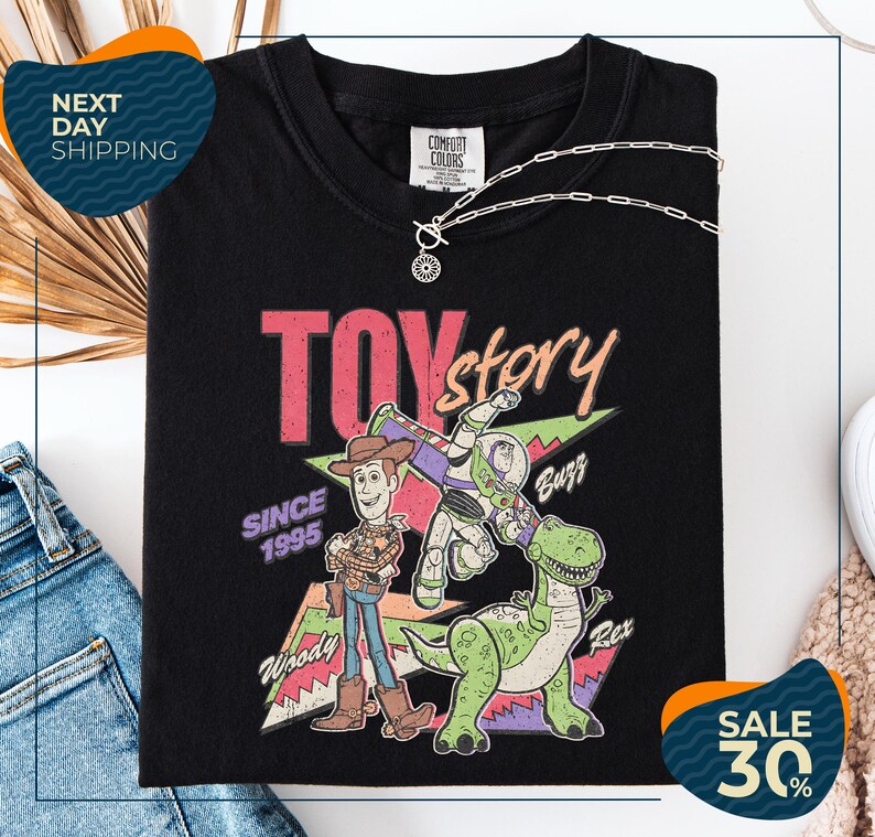 a black t-shirt with a colorful graphic design depicting characters from the Toy Story movie, including Woody and Buzz. The t-shirt is displayed on a white background, and there are jeans and a necklace visible in the foreground.