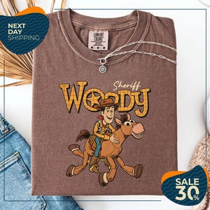 a brown t-shirt with a cartoon image of Woody, a character from the Toy Story franchise, riding a horse. The t-shirt is displayed on a white background, and there are jeans and a sweater visible in the background.