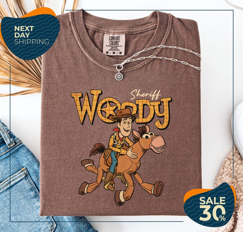 a brown t-shirt with a cartoon image of Woody, a character from the Toy Story franchise, riding a horse. The t-shirt is displayed on a white background, and there are jeans and a sweater visible in the background.