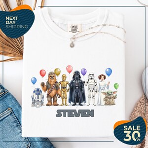 A white t-shirt with a graphic design featuring various characters from the Star Wars franchise, including Chewbacca, Darth Vader, Stormtrooper, and Yoda, along with balloons.