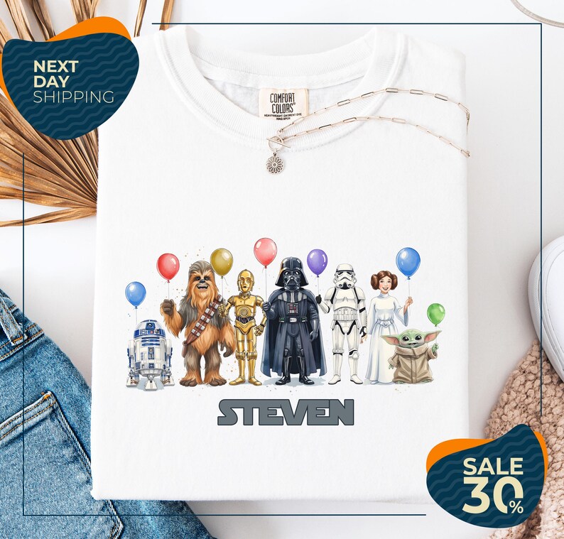 A white t-shirt with a graphic design featuring various characters from the Star Wars franchise, including Chewbacca, Darth Vader, Stormtrooper, and Yoda, along with balloons.