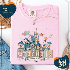a pink t-shirt with a Disney-themed graphic, including Mickey Mouse, Donald Duck, and other Disney characters, along with the text "Graduate from the Tassle to the Castle" and the year "2026". The t-shirt is displayed on a white background, and there are some jeans and a sweater visible in the background.