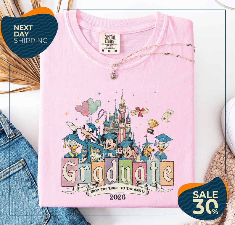 a pink t-shirt with a Disney-themed graphic, including Mickey Mouse, Donald Duck, and other Disney characters, along with the text "Graduate from the Tassle to the Castle" and the year "2026". The t-shirt is displayed on a white background, and there are some jeans and a sweater visible in the background.