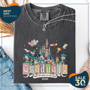 a gray t-shirt with a colorful graphic design depicting Disney characters in a celebratory pose, surrounded by a castle and fireworks. The text on the t-shirt reads "Graduate from the Tassle to the Castle" and "2026". The t-shirt is displayed on a white background, with a pair of jeans and a white object visible in the background.