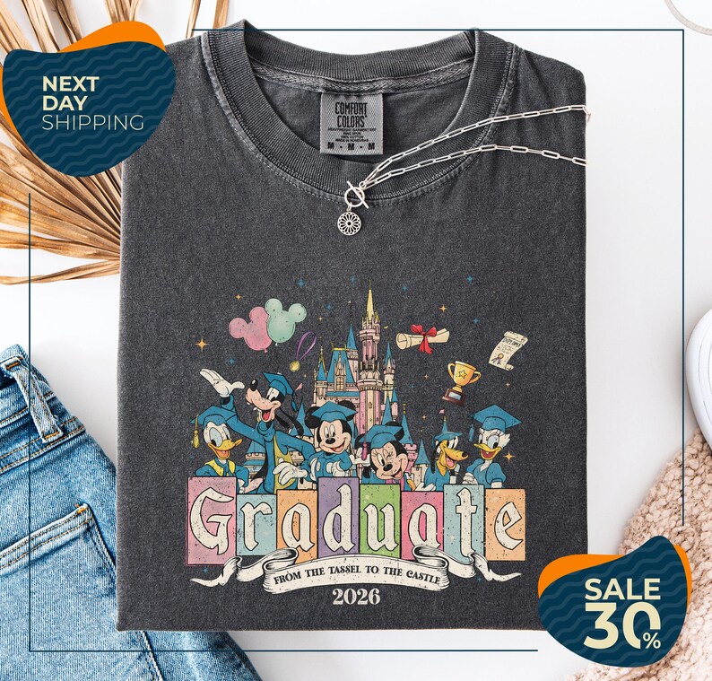 a gray t-shirt with a colorful graphic design depicting Disney characters in a celebratory pose, surrounded by a castle and fireworks. The text on the t-shirt reads "Graduate from the Tassle to the Castle" and "2026". The t-shirt is displayed on a white background, with a pair of jeans and a white object visible in the background.