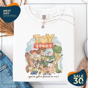 a white t-shirt with a colorful graphic of characters from the Toy Story movie, including Woody, Buzz Lightyear, and other beloved characters. The t-shirt is displayed on a white background, and there are jeans and a sweater visible in the background.
