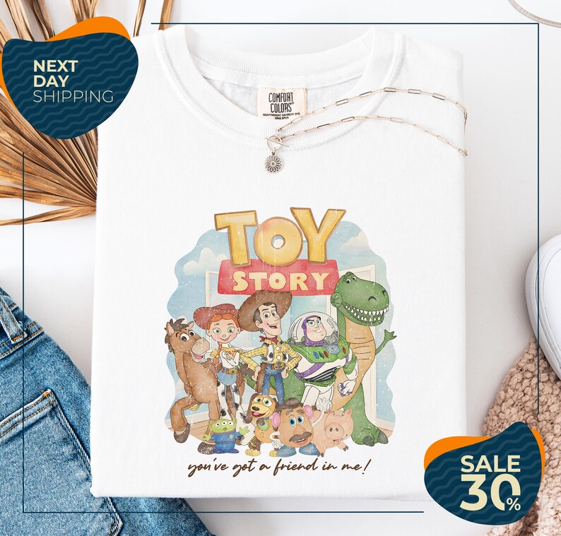 a white t-shirt with a colorful graphic of characters from the Toy Story movie, including Woody, Buzz Lightyear, and other beloved characters. The t-shirt is displayed on a white background, and there are jeans and a sweater visible in the background.